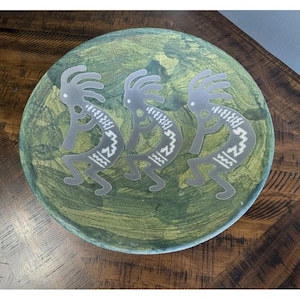 May include: A round, decorative plate with a green and gold painted surface. Three silver Kokopelli figures are affixed to the plate. The Kokopelli figures are in a dancing pose, with a curved body and a headdress.