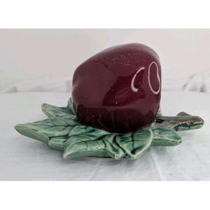 May include: A ceramic salt shaker in the shape of a dark red apple with white accents, resting on a green leaf-shaped dish. The dish has a detailed leaf design. The salt shaker is a decorative kitchen item.