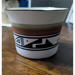 May include: A white ceramic planter with a black rim and a geometric design. The planter features bands of green and brown, and a black and white geometric pattern around the middle. The planter is likely used for indoor plants.