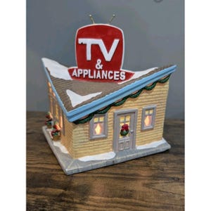 May include: A miniature ceramic building with a red sign that reads "TV & APPLIANCES." The building has a yellow brick facade, a snow-covered roof, and festive decorations. The windows and door are lit up.