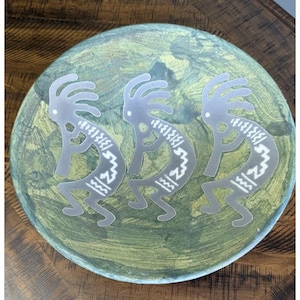May include: A decorative plate with a green and gold painted surface. Three silver Kokopelli figures are arranged across the plate. The Kokopelli figures are in a playing flute pose.