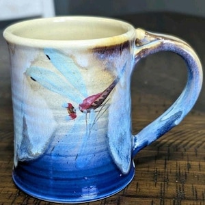 May include: A ceramic mug featuring a hand-painted dragonfly with light blue wings and a red body. The mug has a gradient glaze transitioning from a creamy beige at the rim to a deep, vibrant blue at the base, with a matching blue handle.