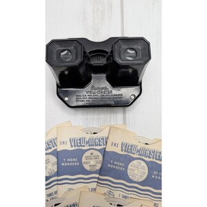 May include: A black plastic Sawyer's View-Master stereoscope viewer with two eyepieces and embossed text on the front. Below the viewer are several paper View-Master reels in blue and white sleeves featuring Grand Teton and Yellowstone National Park.