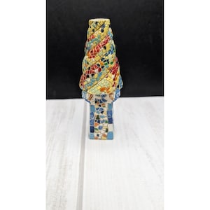 May include: A decorative sculpture featuring a spiral-shaped top covered in a colorful mosaic of yellow, red, blue, and green tiles. The base is rectangular and also decorated with a mosaic pattern of blue, white, orange, and yellow tiles.