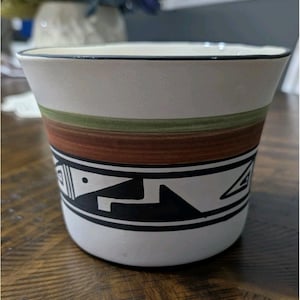 May include: A white ceramic planter with a black rim and a decorative band featuring geometric shapes in black. The planter has bands of green and brown, and is suitable for indoor or outdoor use.