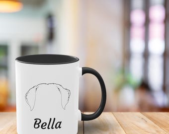 Peut inclure: Mug en céramique blanche avec intérieur et anse noirs. La tasse présente un dessin au trait d'oreilles de chien et le nom "Bella" en noir, dans une police simple. La tasse est posée sur une surface en bois.