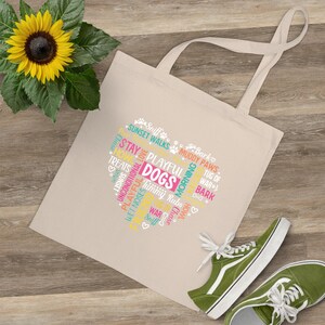 Op de afbeelding: Een beige canvas draagtas met een hartvormig ontwerp gevuld met honden gerelateerde woorden zoals "playful" en "treats". Het woord "DOGS" staat prominent afgebeeld. Een gele zonnebloem en groene sneakers zijn ook zichtbaar.