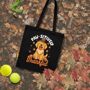 Op de afbeelding: Zwarte draagtas met een lachende golden retriever met een pelgrimshoed, omringd door pompoenen en herfstbladeren. De tekst luidt "Paw-sitively Thankful". Twee gele tennisballen zijn zichtbaar.