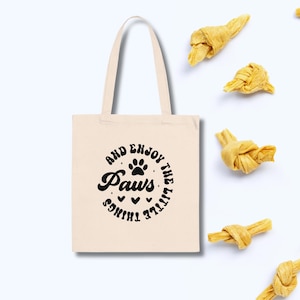 May include: A cream-coloured tote bag with black text that reads "AND ENJOY THE LITTLE THINGS PAWS". The bag has a paw print design and several dog treats are scattered around it.