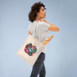Op de afbeelding: Crèmekleurige draagtas met een kleurrijk pootafdrukontwerp. De pootafdruk heeft roze, blauwe en groene accenten. De tas wordt over de schouder gedragen en bevat een brood.