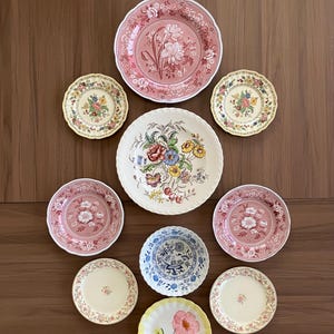 May include: A collection of decorative plates with floral designs. The plates vary in size and color, including pink, yellow, and blue. Some plates feature detailed floral patterns, while others have simpler designs. The plates are arranged on a wooden surface.
