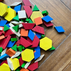May include: A collection of colorful wooden geometric shapes, including triangles, squares, hexagons, and diamonds, in various colors like red, yellow, green, blue, and orange. The shapes are scattered on a wooden surface.