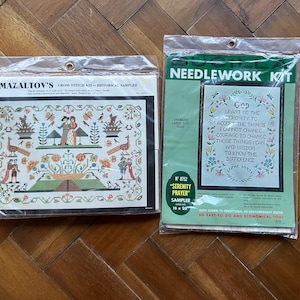 May include: Two cross-stitch kits in plastic packaging. One is a historical sampler with a colorful design and the text "MAZALTOV'S CROSS STITCH KIT". The other is a "SERENITY PRAYER" sampler, 16 x 20 inches, with a floral border and text.