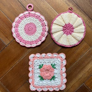 May include: Three handmade crochet pot holders in shades of pink and white. Two are round with a loop for hanging, and one is square with a floral design. The pot holders are made with a textured stitch.