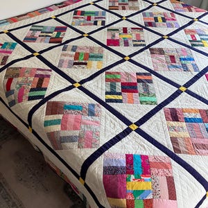 May include: A patchwork quilt featuring a grid of square blocks made from various multi-colored fabric scraps. The blocks are separated by dark blue sashing with small yellow corner squares, all set against a cream-colored background.