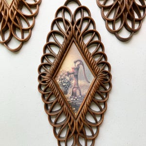 May include: Set of three vintage wall art pieces. Each piece features a diamond-shaped frame with a woven design. The frames surround a picture of a water pump and flowers. The frames are a medium brown color.