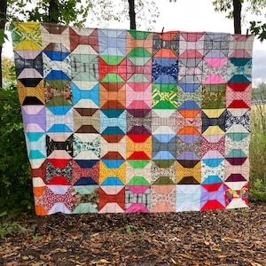 May include: A handmade quilt with a repeating bow tie pattern. The quilt features a variety of colorful fabrics, including floral, geometric, and solid patterns. The quilt is displayed outdoors, hanging between two trees.