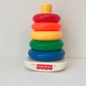 May include: A Fisher-Price stacking toy with a white base featuring the text U.S.A. and 1050. The toy consists of a central post holding five plastic rings in descending order of size, colored blue, green, yellow, orange, and red, topped with a yellow cap.
