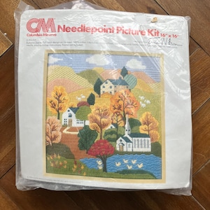 May include: A sealed needlepoint picture kit featuring an autumn scene with houses, trees, and a lake. The kit is 16" x 16" and includes a full-color design on 14-mesh canvas. The brand is Columbia-Minerva.