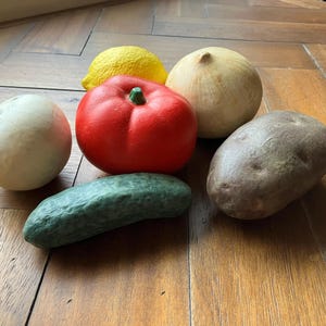 May include: A collection of colorful, artificial produce including a red tomato, yellow lemon, green cucumber, brown potato, and two white onions. These decorative items are arranged on a wooden surface.