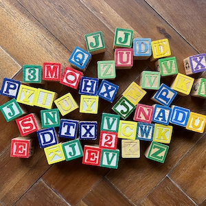 May include: A collection of colorful wooden alphabet blocks scattered on a wooden surface. Each block features a letter or number in a bold font, with various colors like red, green, blue, and yellow. The blocks are a classic educational toy.