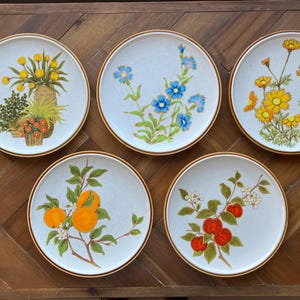 May include: Set of five white ceramic dinner plates with gold trim. Each plate features a different floral design with yellow, blue, and orange flowers.