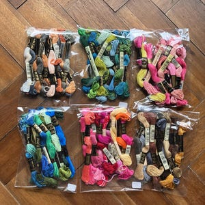 May include: Six clear plastic bags filled with embroidery floss in various colors. The floss colors include shades of pink, green, blue, orange, and brown. Each bag contains multiple spools of thread, perfect for crafting and embroidery projects.