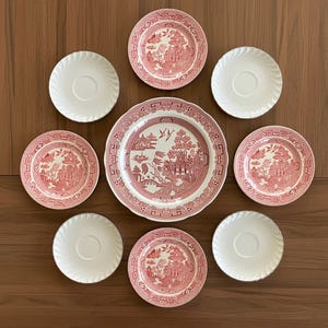 May include: A collection of vintage china featuring a red and white Willow pattern. The set includes a large dinner plate, several smaller plates, and saucers. The plates are arranged on a brown wooden surface.