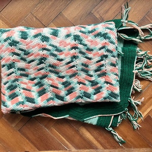 May include: A crocheted blanket with a green and pink chevron pattern. The blanket has a green border and fringe on all sides.