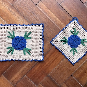 May include: Two square crochet potholders made of off-white yarn with a central blue flower motif and green leaves. Each piece features a blue crocheted border with a small loop for hanging. One is oriented squarely, while the other is rotated.