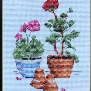 May include: Cross stitch pattern featuring two potted geraniums, one with red flowers and the other with pink flowers. The pattern includes the text "Geraniums" and "Punto de Cruz Comptado".