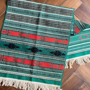 May include: A green, red, and black striped table runner with fringe on the ends. The runner has a geometric pattern.