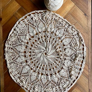 May include: A cream-colored, circular doily with an intricate crochet pattern featuring floral and geometric designs. A ball of white yarn sits above the doily. The doily is placed on a wooden surface.