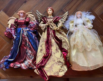 May include: Three decorative angel figurines with detailed features. The angels have fabric gowns in red, blue, gold, and white. Each angel has wings and a serene expression. The angels are displayed on a wooden surface.