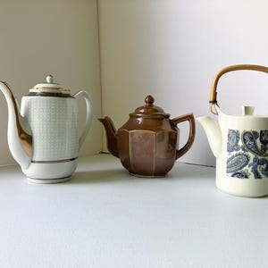 May include: Three teapots of varying styles. The first is white with gold trim and a geometric pattern. The second is brown and hexagonal. The third is white with a blue paisley design and a bamboo handle.