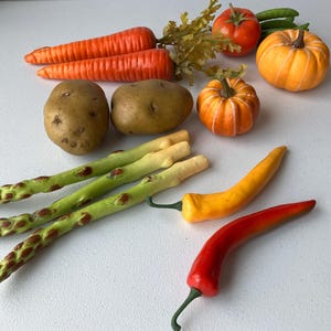 May include: A collection of assorted vegetables, including two orange carrots, two brown potatoes, four green asparagus spears, one yellow and orange pepper, one orange pumpkin, and one red chili pepper.