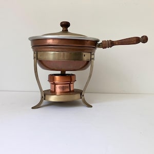 May include: A copper fondue pot with a brass stand and burner. The pot features a silver-toned rim, a wooden handle, and a matching wooden knob on the lid. The brass stand has three curved legs and a circular base holding a small copper burner.