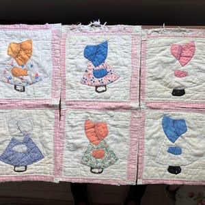 May include: Six square quilt blocks featuring appliquéd figures wearing bonnets. Each block has a white background with a pink checkered border. The figures are dressed in various patterned fabrics, including floral and solid colors like orange, blue, and pink.