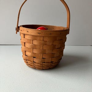 Op de afbeelding: Een kleine, bruine geweven mand met een houten handvat. De mand is leeg, maar er zit een rode appel in.