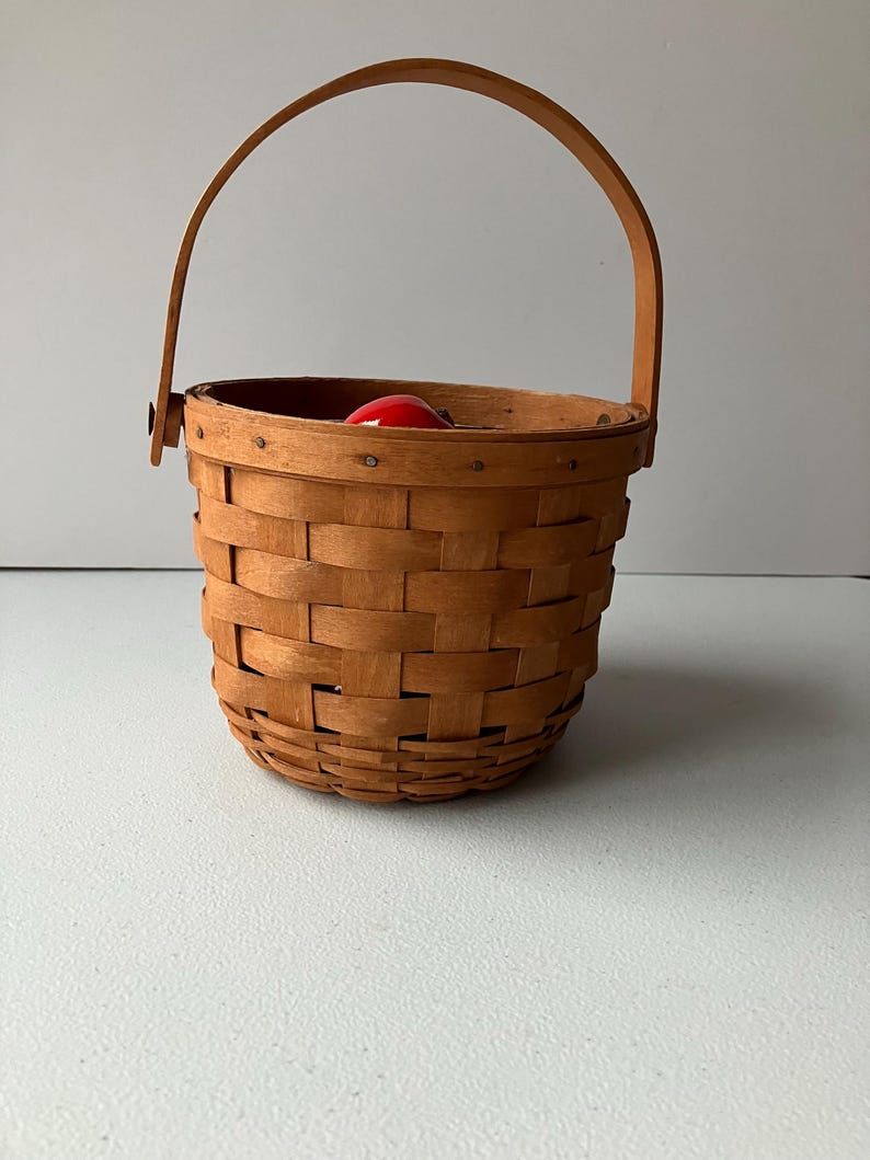 Op de afbeelding: Een kleine, bruine geweven mand met een houten handvat. De mand is leeg, maar er zit een rode appel in.