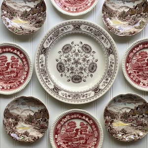May include: A collection of vintage ceramic plates with various designs. The plates feature a mix of colors, including red, brown, and white. Some plates have a landscape scene, while others have floral patterns. The plates are arranged on a white surface.