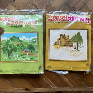 May include: Two vintage Sunset needlepoint kits. One kit features a green package with a landscape scene of a red barn, trees, and rolling hills. The other kit has a gold package with a house and tree scene. Both kits are sealed in plastic.