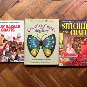 May include: Three vintage craft books titled "Easy Bazaar Crafts", "Creative Crafts and Stitchery", and "Stitchery and Crafts" by Better Homes and Gardens. The books are on a wooden surface. The middle book features a butterfly design.