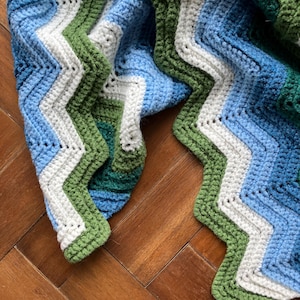 May include: A handmade crochet blanket with a chevron pattern. The blanket features a combination of blue, white, and green yarn. The edges are finished with a green border. The blanket is folded on a wooden surface.