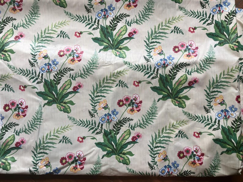 May include: Cream-coloured fabric with a floral pattern. The design includes bouquets of pink, blue, and yellow flowers, interspersed with green fern leaves, set against a backdrop of vertical stripes. The fabric has a sheen.