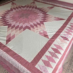 May include: A large quilt featuring a central star pattern composed of various shades of pink and white fabric. The design incorporates geometric diamond shapes and solid white squares, finished with a wide pink border and a floral patterned edge.