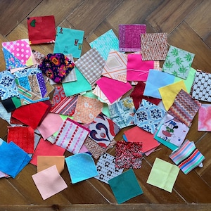 May include: A collection of colorful fabric squares in various patterns and colors, including pink, blue, red, and green. The squares are arranged on a wooden surface, showcasing a variety of textures and designs, suitable for quilting or crafting projects.