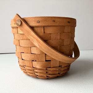 Op de afbeelding: Een kleine, bruine geweven mand met een houten handvat. De mand is gemaakt van natuurlijk hout en heeft een eenvoudig, rustiek ontwerp.
