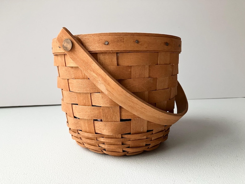 Op de afbeelding: Een kleine, bruine geweven mand met een houten handvat. De mand is gemaakt van natuurlijk hout en heeft een eenvoudig, rustiek ontwerp.