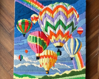 May include: A colorful needlepoint wall hanging featuring a scene of hot air balloons floating in the sky with a rainbow in the background. The balloons are various colors, including red, green, yellow, orange, and purple. The sky is blue with white clouds. The background is a green field.